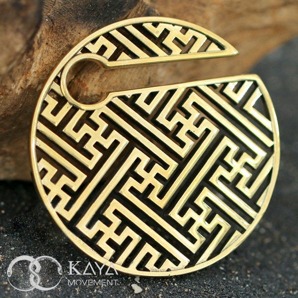 The SAYL Sayagata Disc Ear Weights in Gold by Kaya Movement showcase a round, gold-colored pendant adorned with an intricate geometric pattern of interconnected lines and shapes, reminiscent of Massif Sayagata ear weights. The design is partially open at the top, and this exquisite piece of body jewelry is labeled with "Kaya Movement" and a logo in the lower left corner.